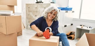 Middle age grey-haired woman smiling happy packing kitchen cardboard box at new home.